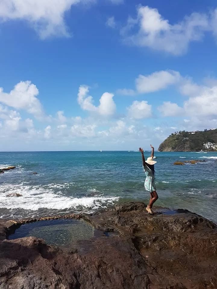 Paula standing on rocks at the shore, arms raised, open sky behind