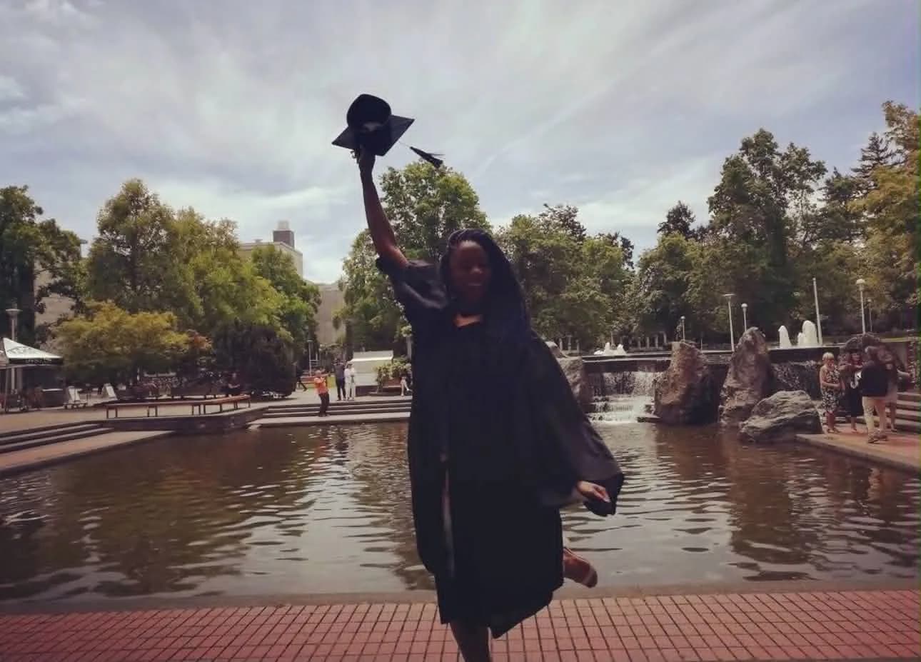 Paula in a graduation gown by a fountain, raising her cap in the air